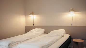 Una cama bien hecha con sábanas blancas, un taburete de madera y dos lámparas fijadas a la pared.