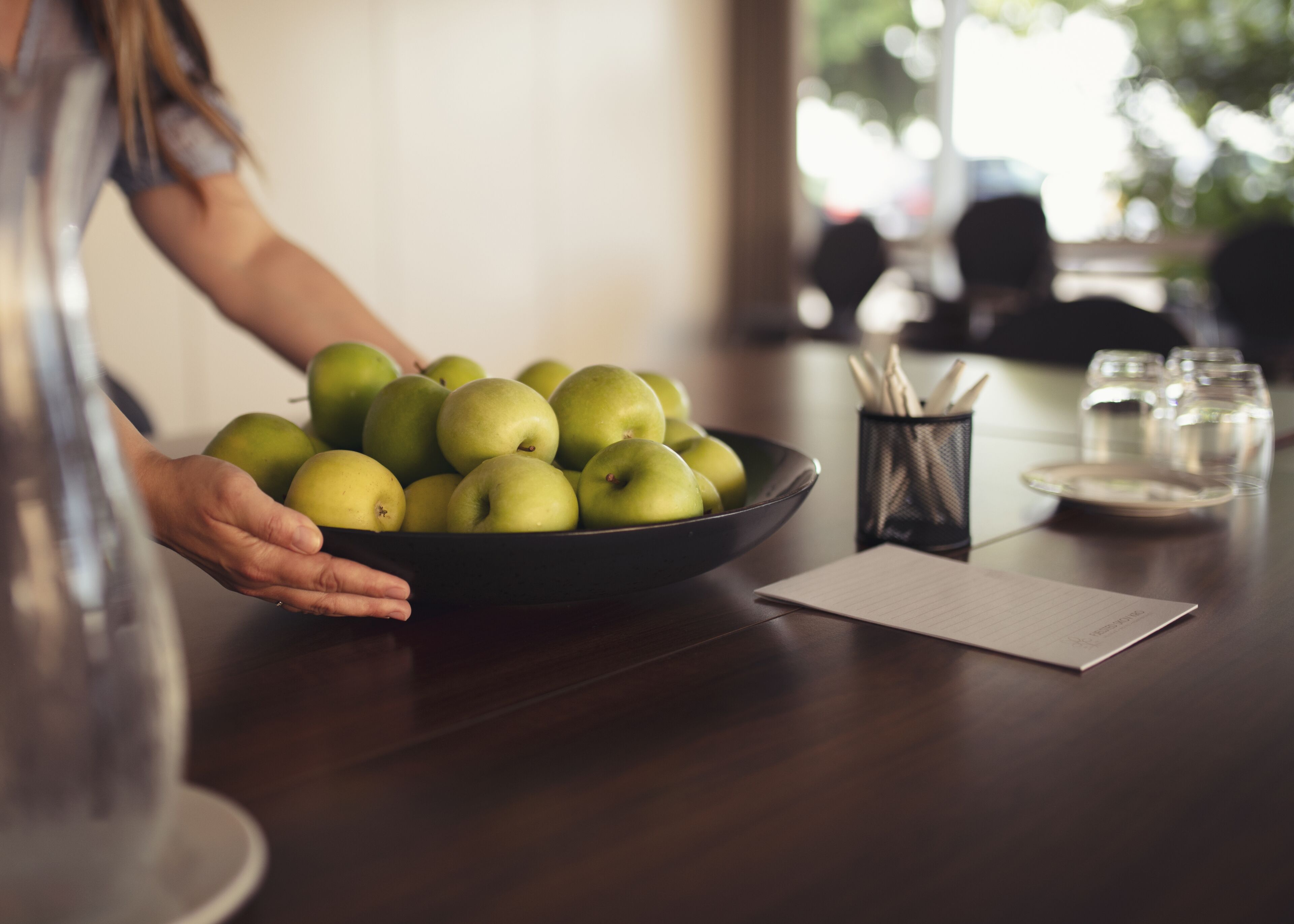 Una persona sosteniendo un bol de manzanas verdes sobre una mesa de madera, con un vaso de agua, un bloc de notas y un portalápices.