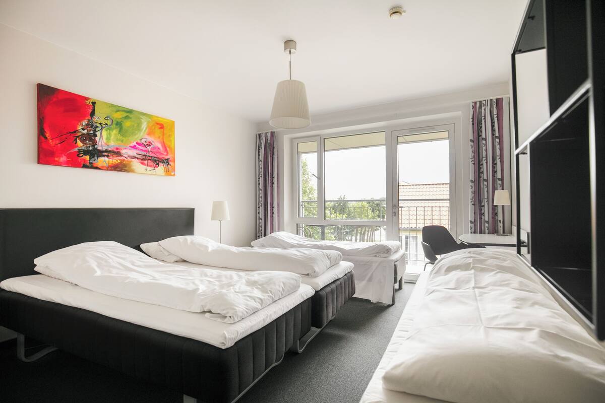 A hotel room with two beds, a large window, a painting, and a ceiling light.