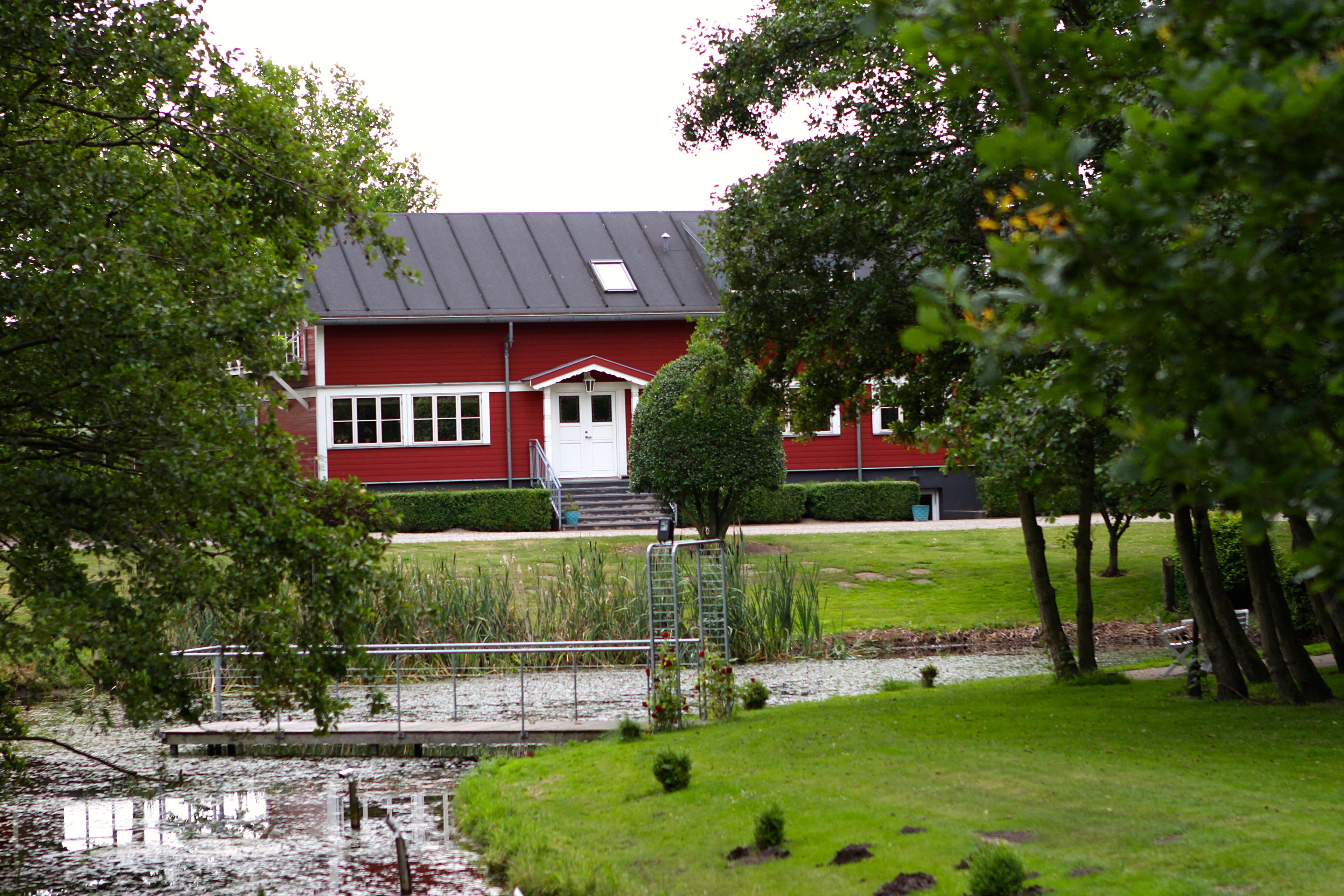 En röd byggnad med en vit dörr och vita fönster, omgiven av träd och en liten damm.