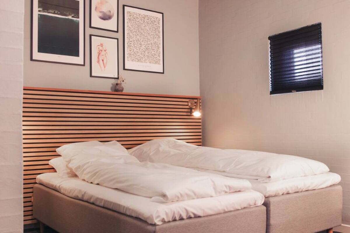 A neatly made bed with white linens in a room with a slatted headboard, a small window with blinds, and framed artwork on the wall.