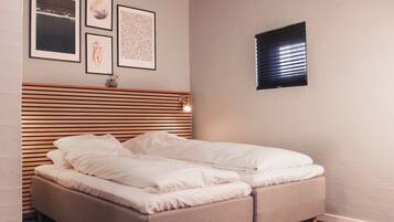 Een net opgemaakt bed met witte lakens in een kamer met een latten hoofdbord, een klein raam met zonwering en kunstwerken in een lijst aan de muur.