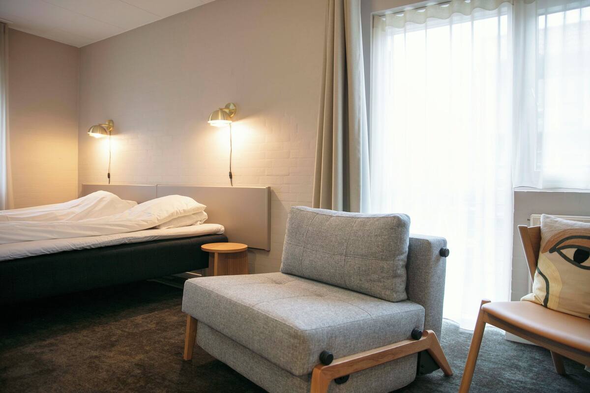 A hotel room with a bed, a grey armchair, a small wooden stool, and a window with sheer curtains.