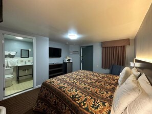 Quarto de hotel com uma cama grande, uma escrivaninha, uma cadeira, uma televisão e um banheiro visível através de uma porta aberta.