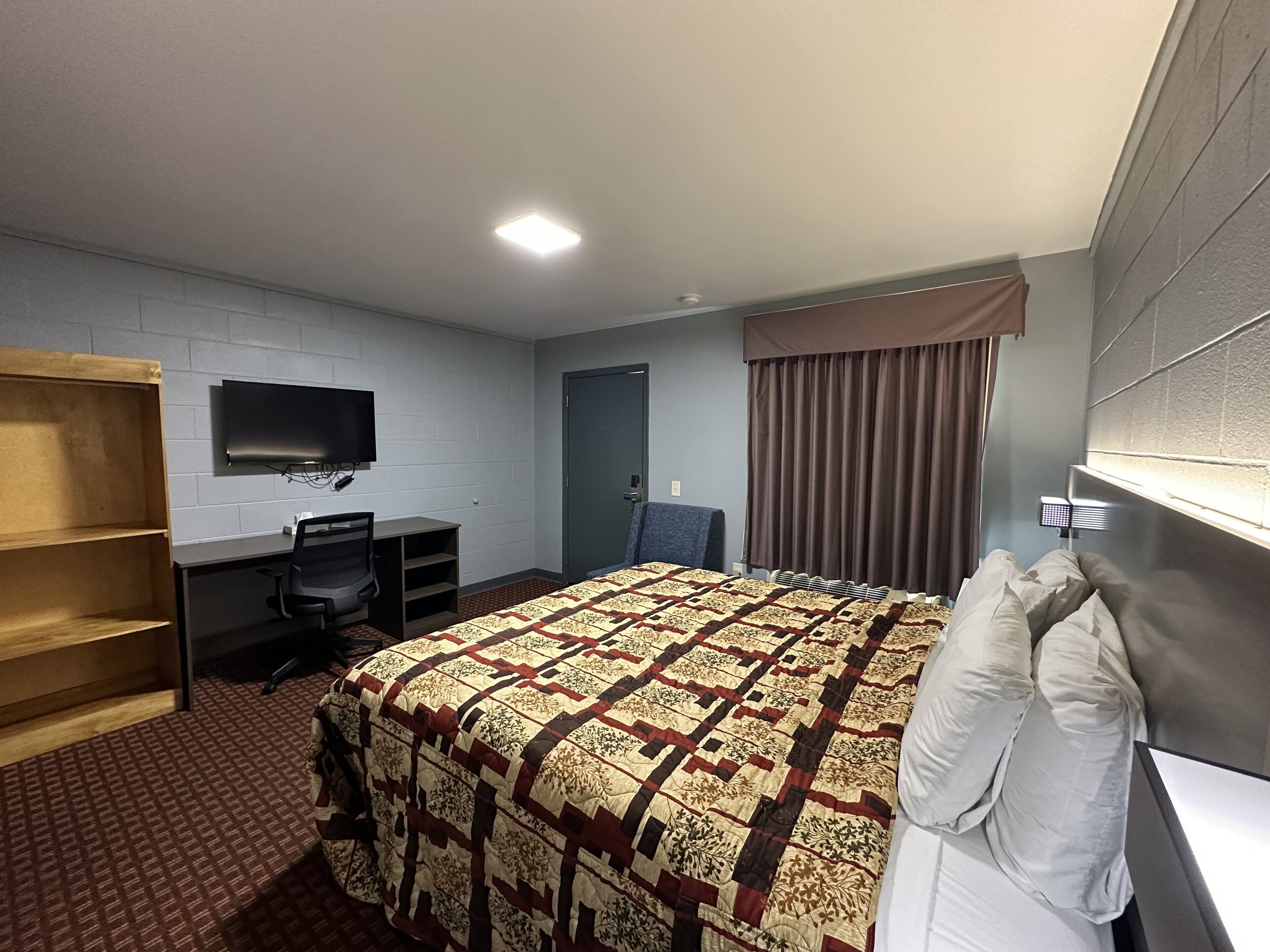 Ein Hotelzimmer mit Bett, Schreibtisch, Sessel, Fernseher und einem Fenster mit Vorhängen.