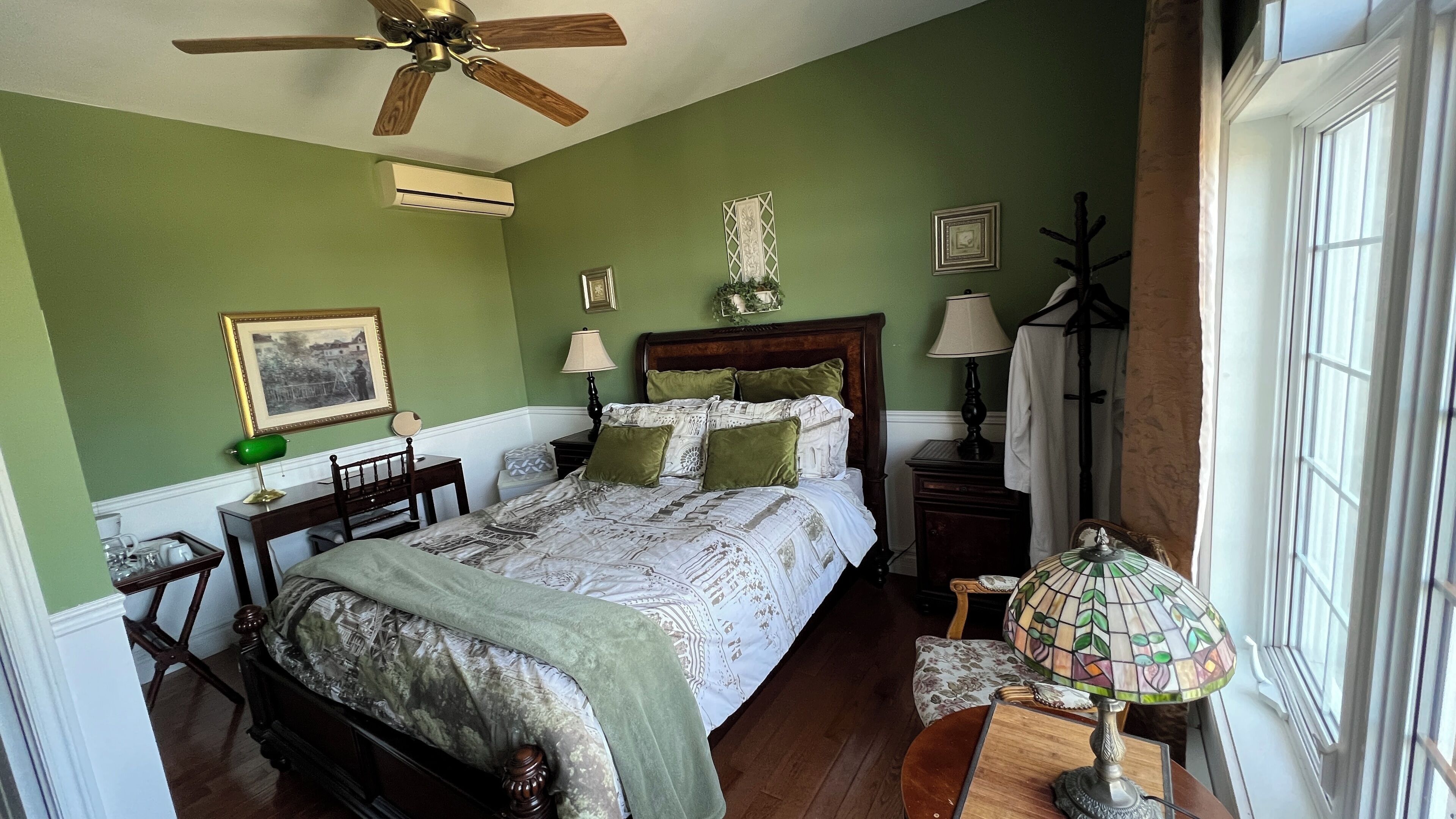 Un dormitorio con cama de madera, paredes verdes, ventilador de techo, equipo de aire acondicionado, mesita de noche con lámpara, un cuadro enmarcado y una ventana con cortinas.