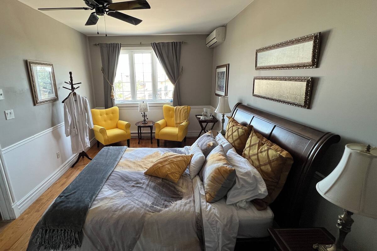 A hotel room with a bed, a yellow armchair, a small table with a lamp, and a window with curtains.