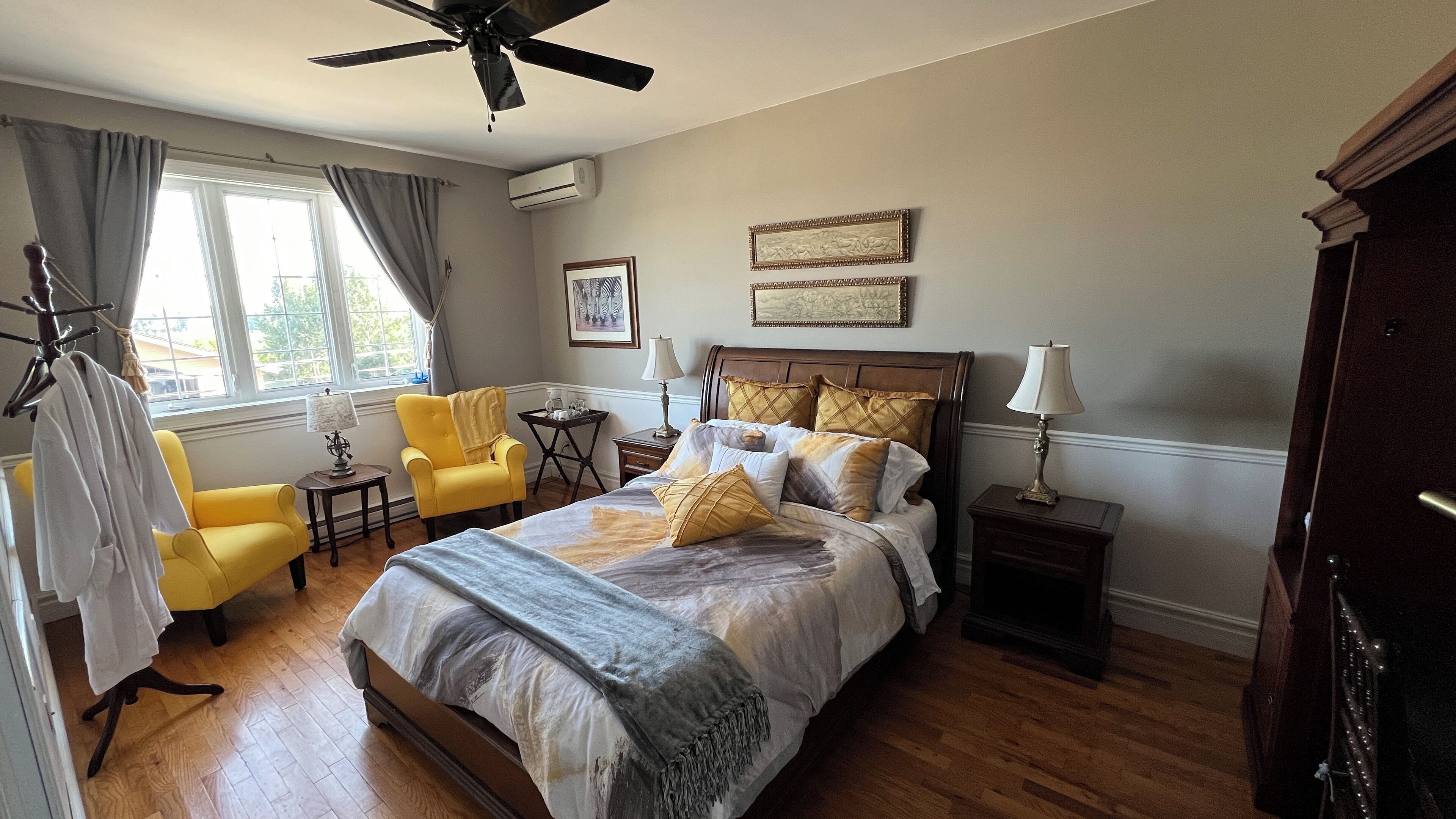 A well-lit bedroom with a large bed, a ceiling fan, a window with curtains, a chair, a small table with a lamp, and a nightstand with a lamp.