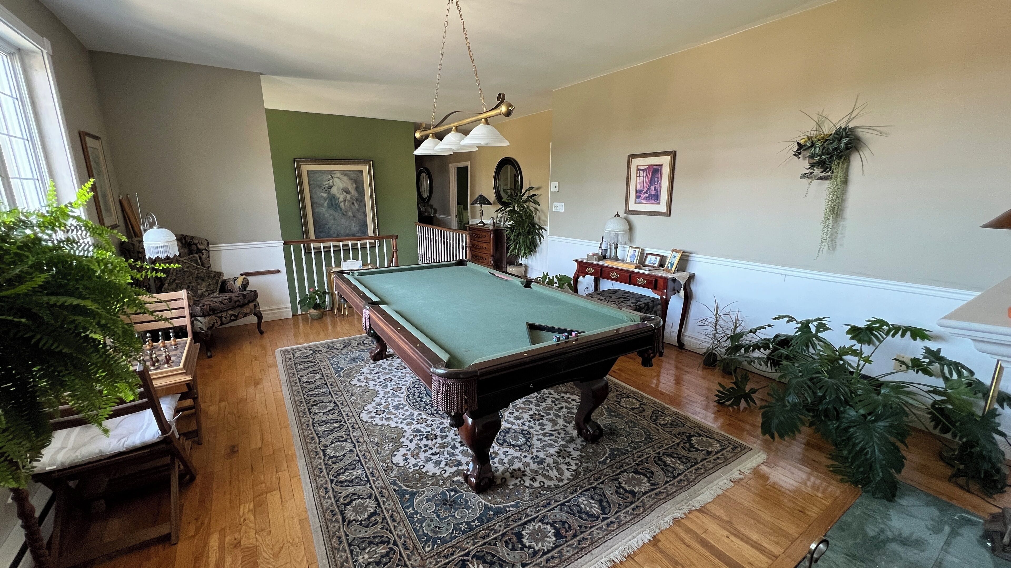Une table de billard dans une pièce au plancher en bois, aux murs verts et agrémentée de diverses plantes.