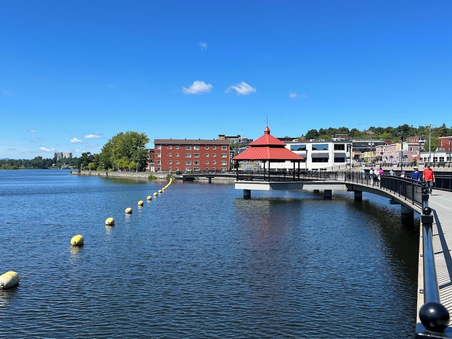 A waterfront promenade with a red-roofed pavilion, a bridge, and buildings in the background.