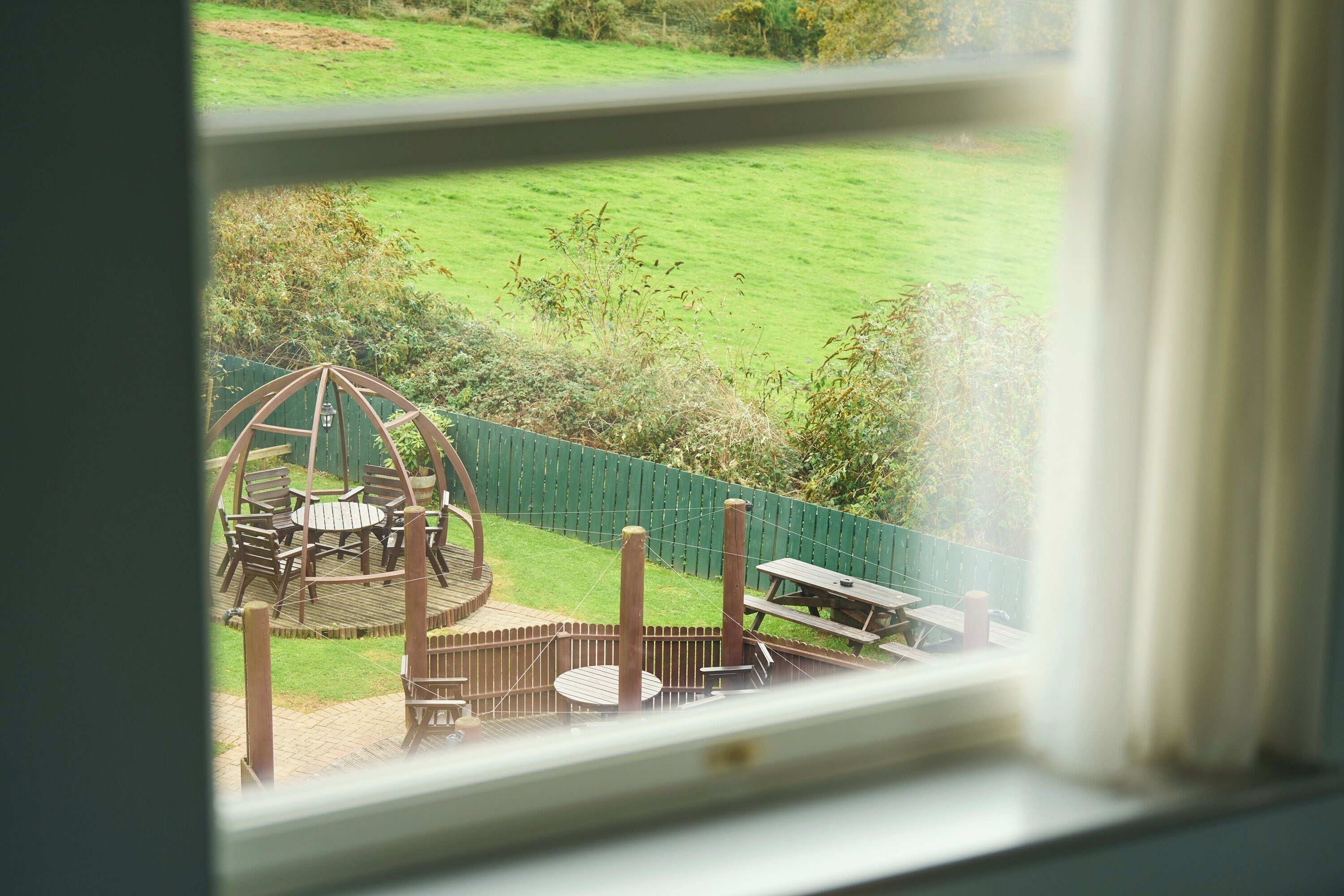 A view through a window of a garden with a wooden deck, a table, and chairs.