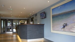 Uma área de recepção com piso de madeira, balcão de atendimento, um quadro decorativo com uma praia e uma televisão na parede.