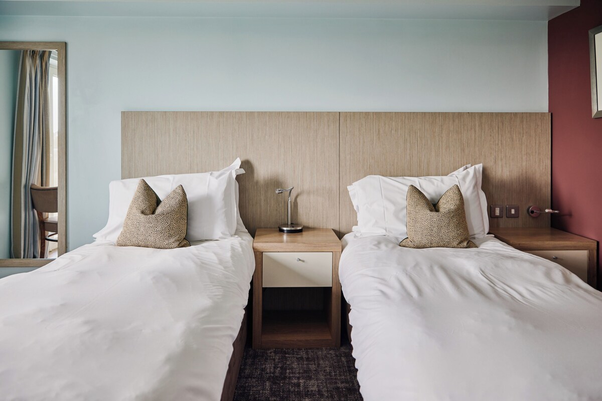 Una habitación de hotel con dos camas individuales, una mesita de noche con lámpara y un cabecero de madera.