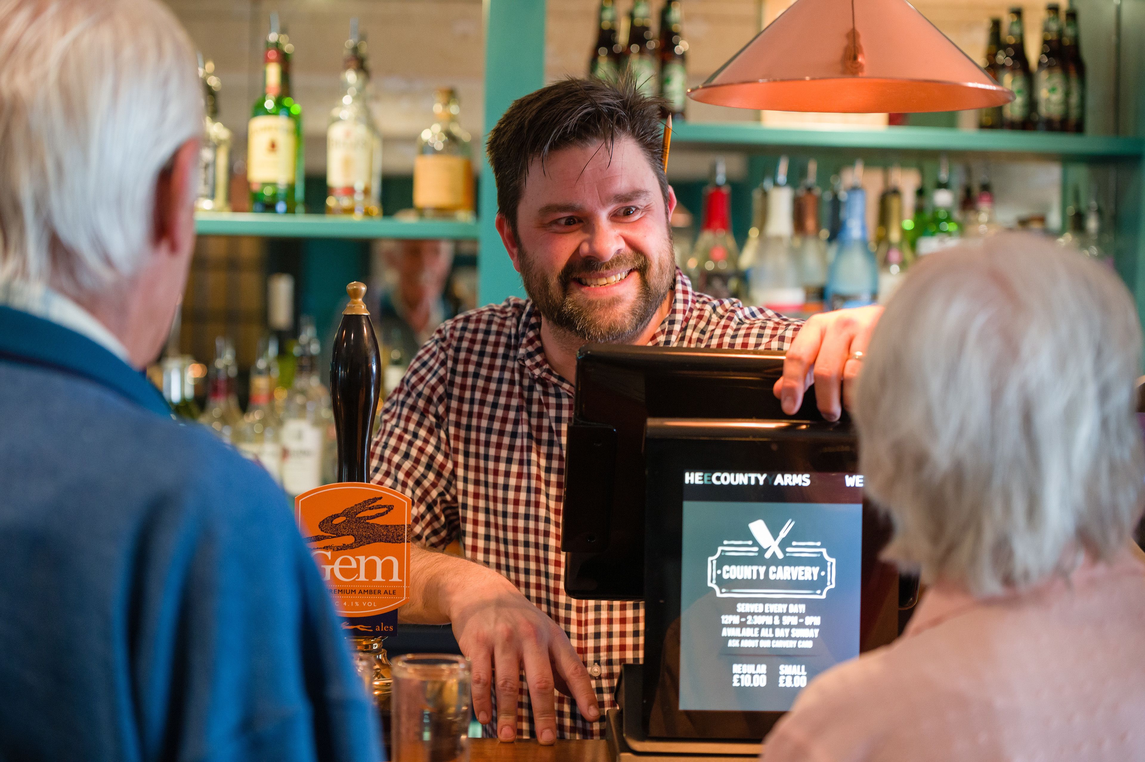 A bar setting with a person holding a menu, various bottles on shelves, and a sign indicating a carvery service.