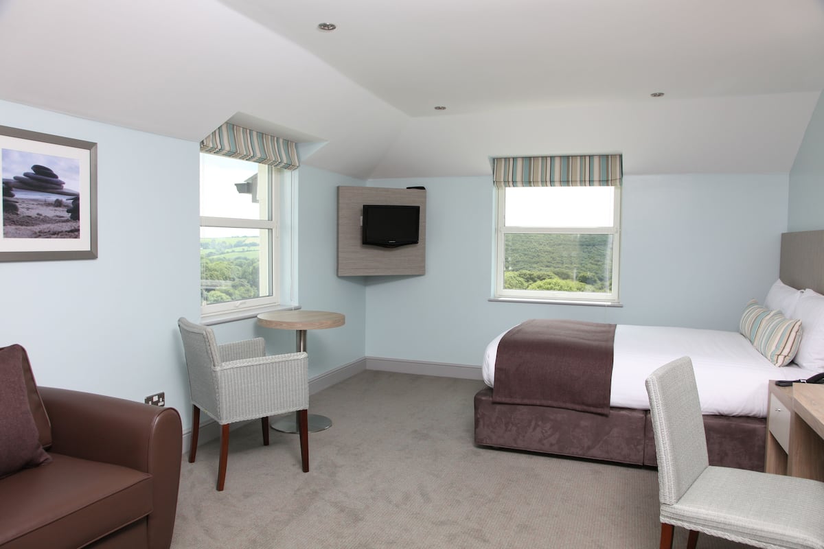 A bedroom with a bed, a chair, a small table, a TV, and a window with a view of greenery.
