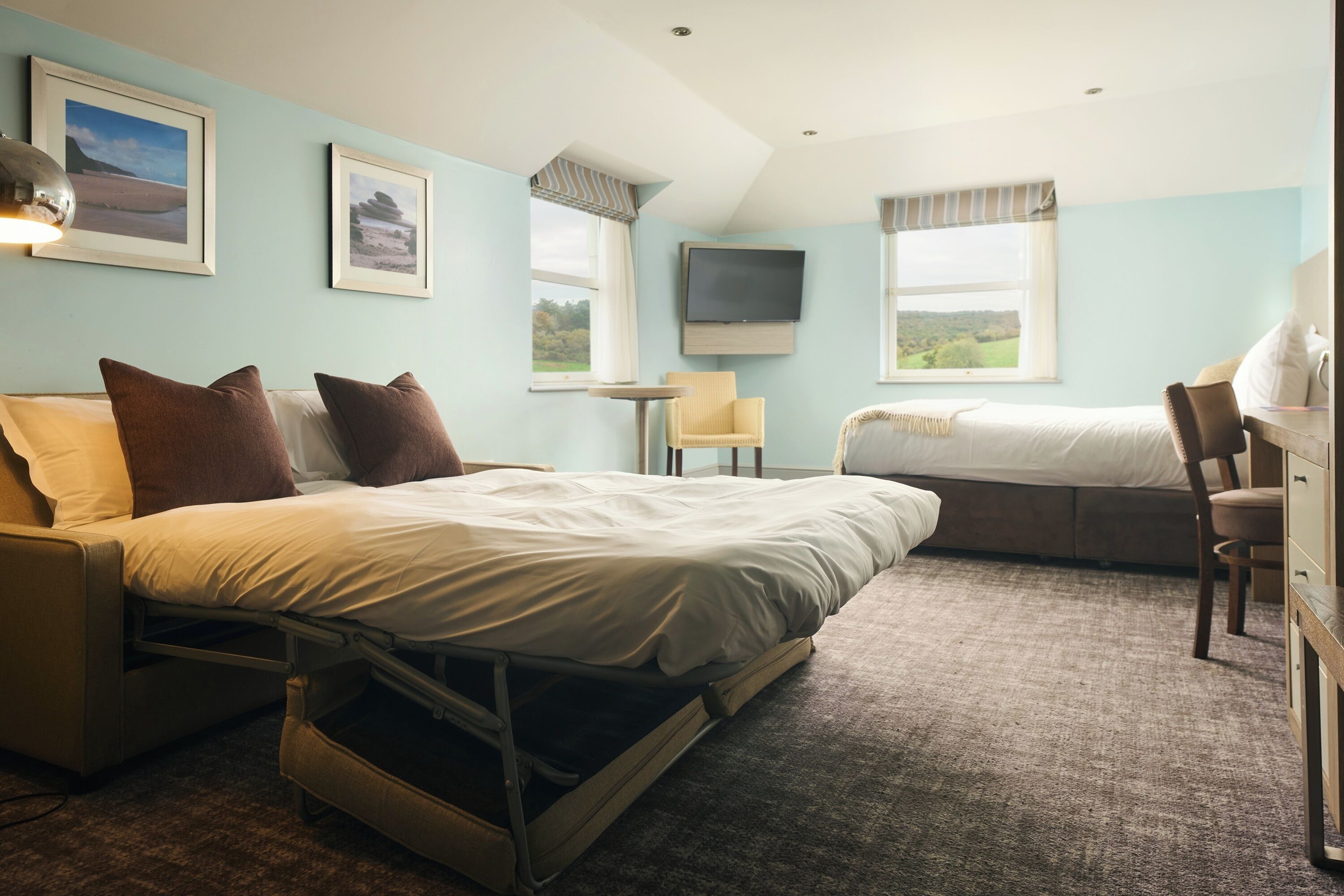 Ein Hotelzimmer mit einem Bett, einem Schreibtisch, einem Sessel, einem Fernseher und zwei Fenstern.