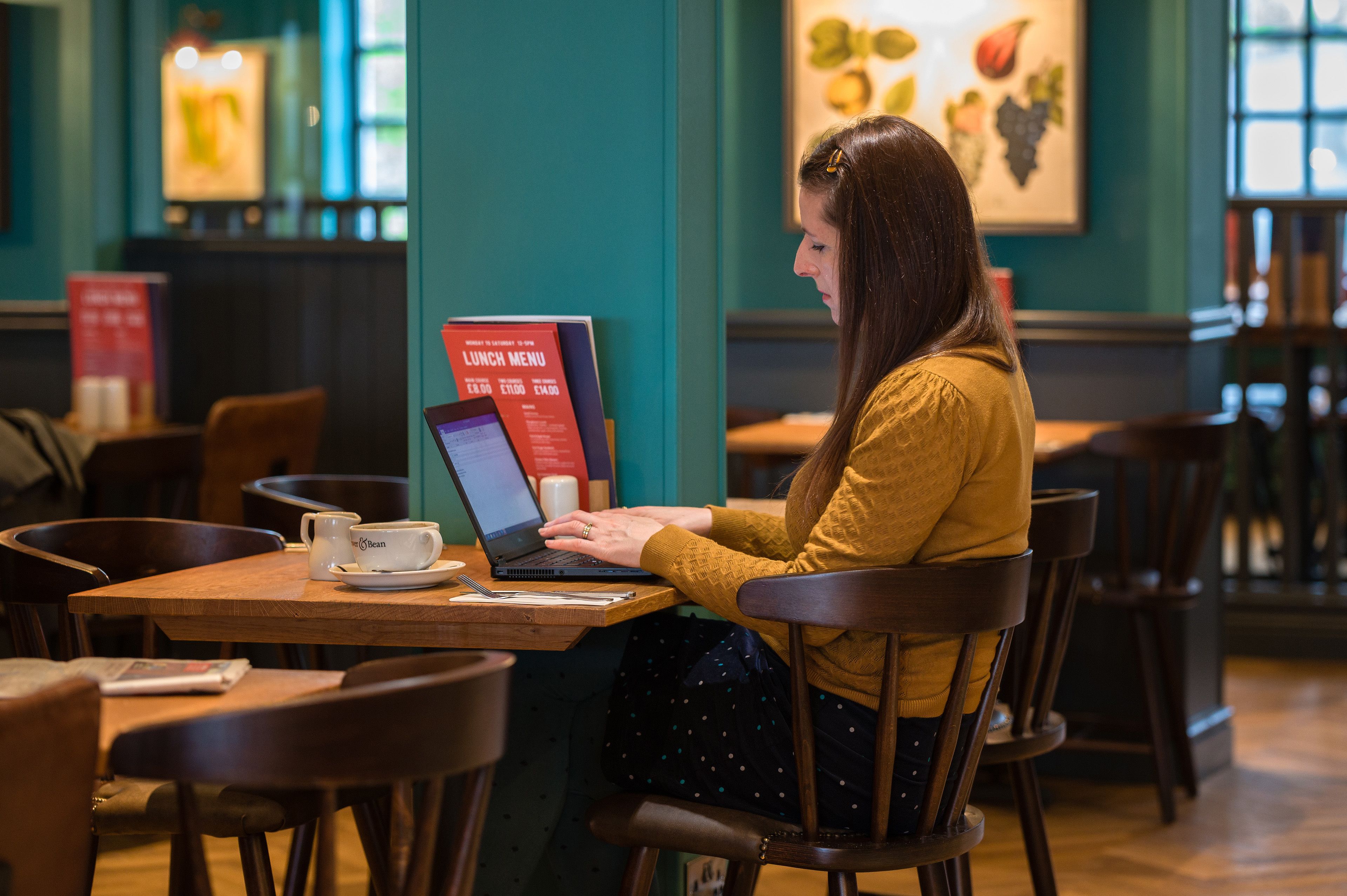 Une personne assise à une table en bois, devant un ordinateur portable, un menu et une tasse.