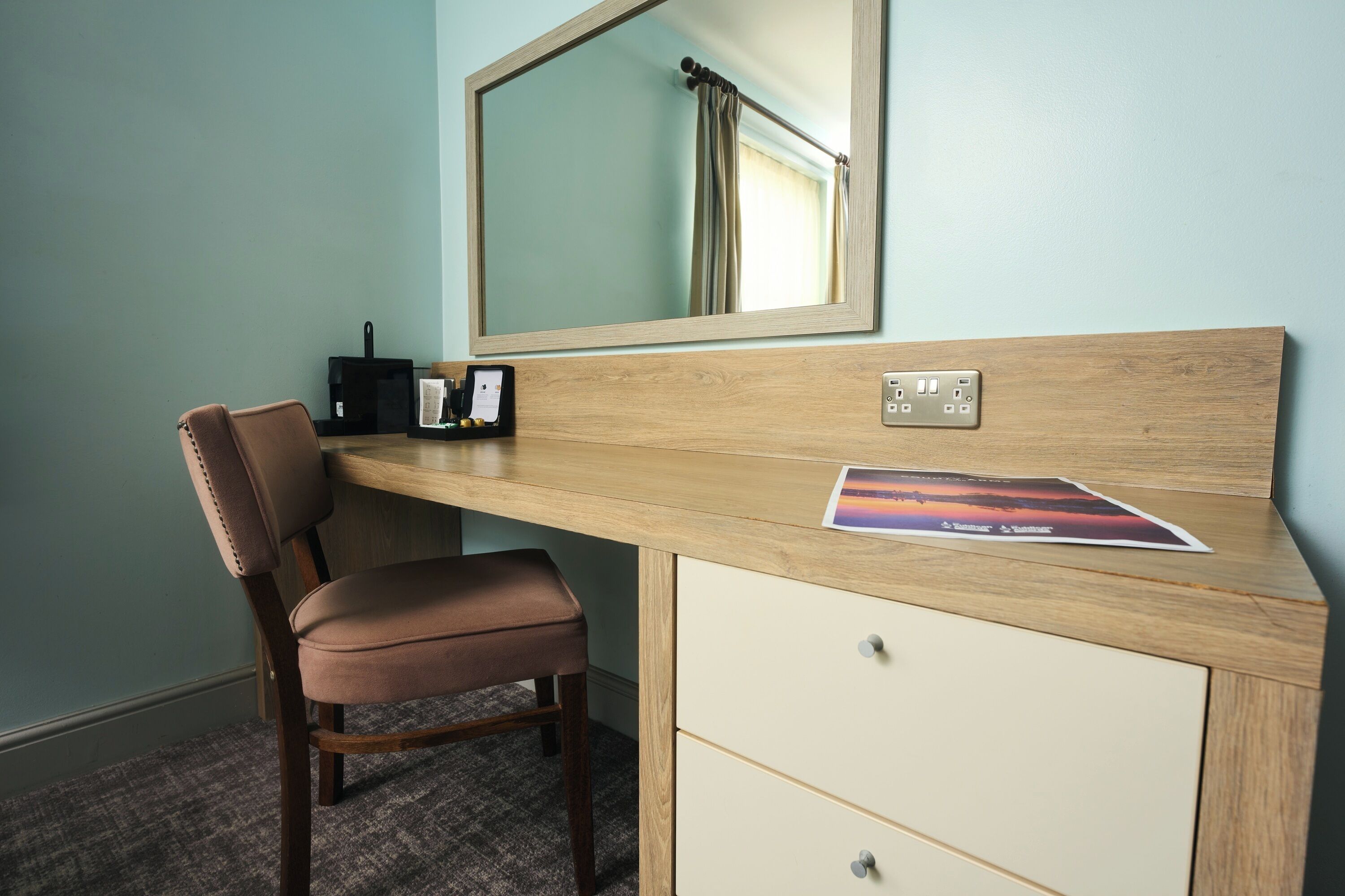 A hotel room with a wooden desk, a chair, a mirror, a small black radio, and a picture frame.