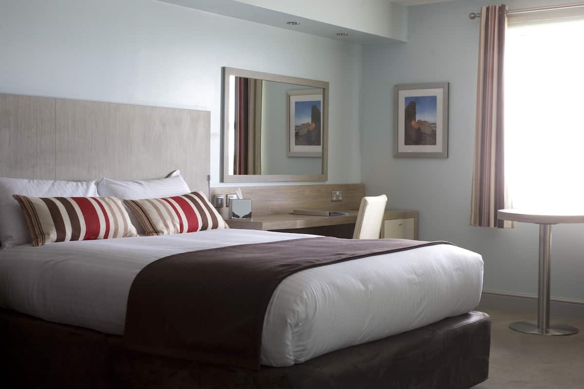 A hotel room with a large bed, a bedside table, a mirror, and a window with curtains.