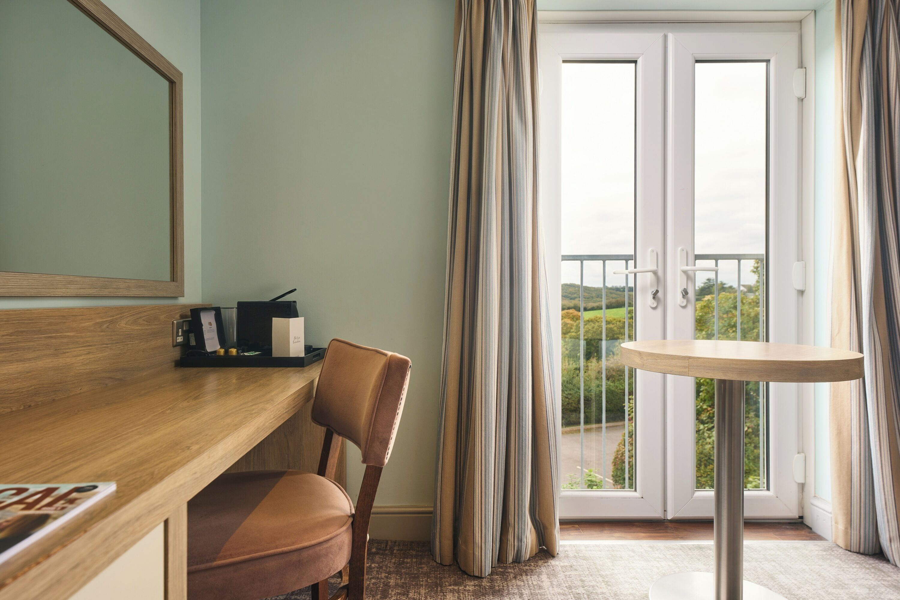 Habitación de hotel con escritorio de madera, una silla, una mesita y vistas a un balcón a través de una puerta de vidrio.