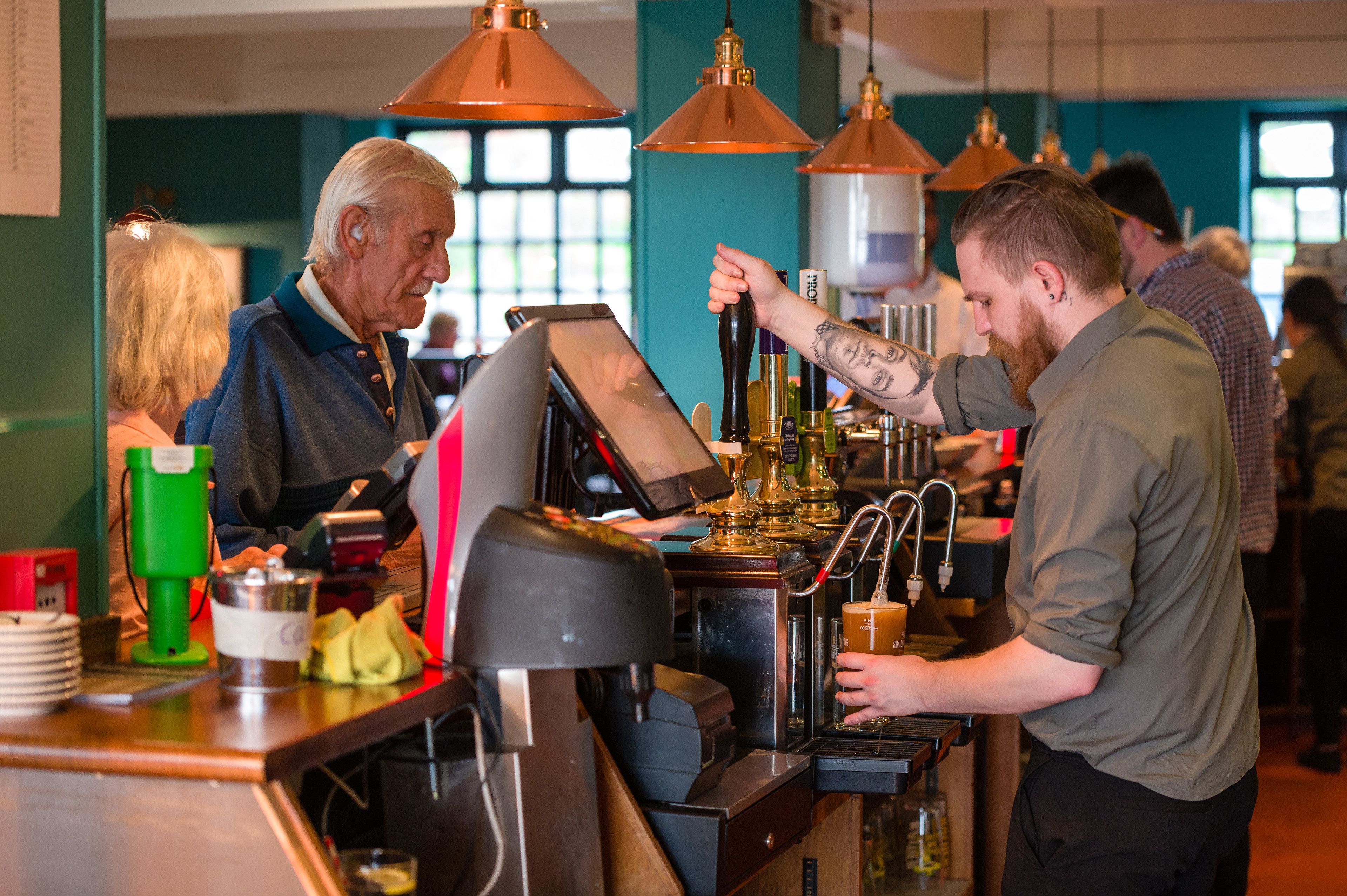A barista preparing a drink at a coffee machine in a busy cafe with other patrons in the background.