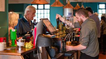 A barista preparing a drink at a coffee machine in a busy cafe with other patrons in the background.