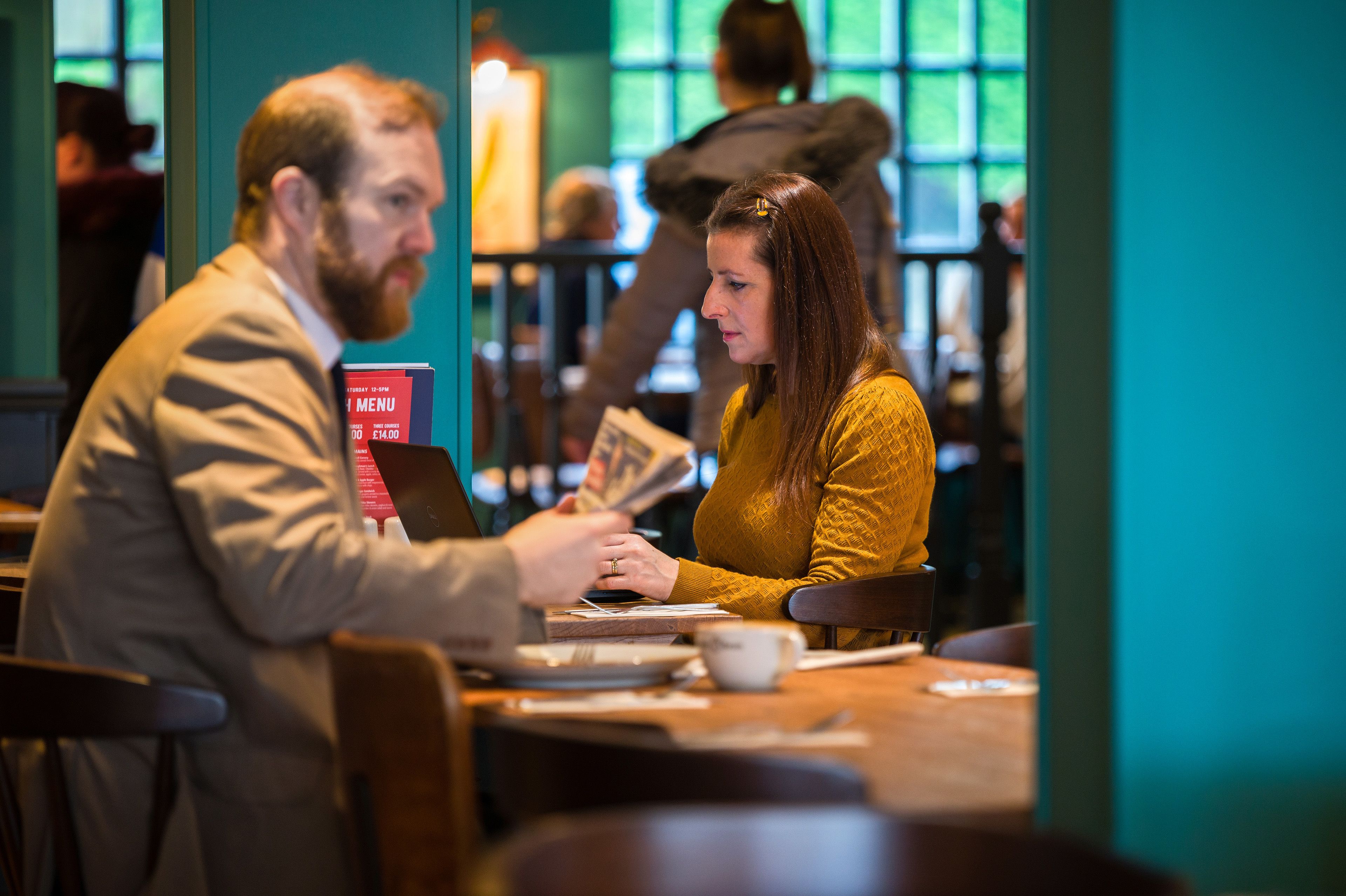 A person handing over a menu to another person at a table in a cafe.
