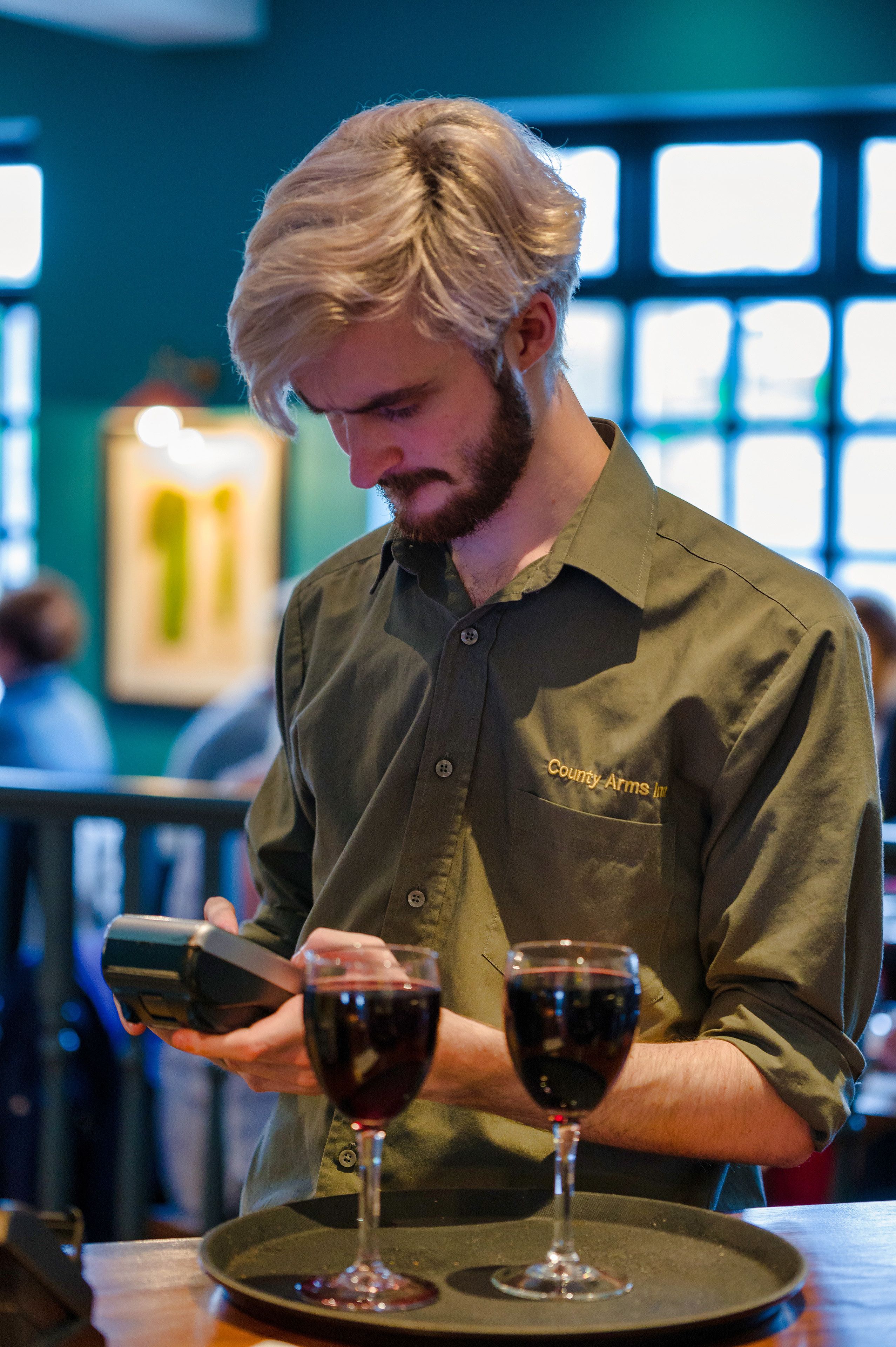 A person in a green shirt with "County Arms Inn" on it, pouring wine into a glass.