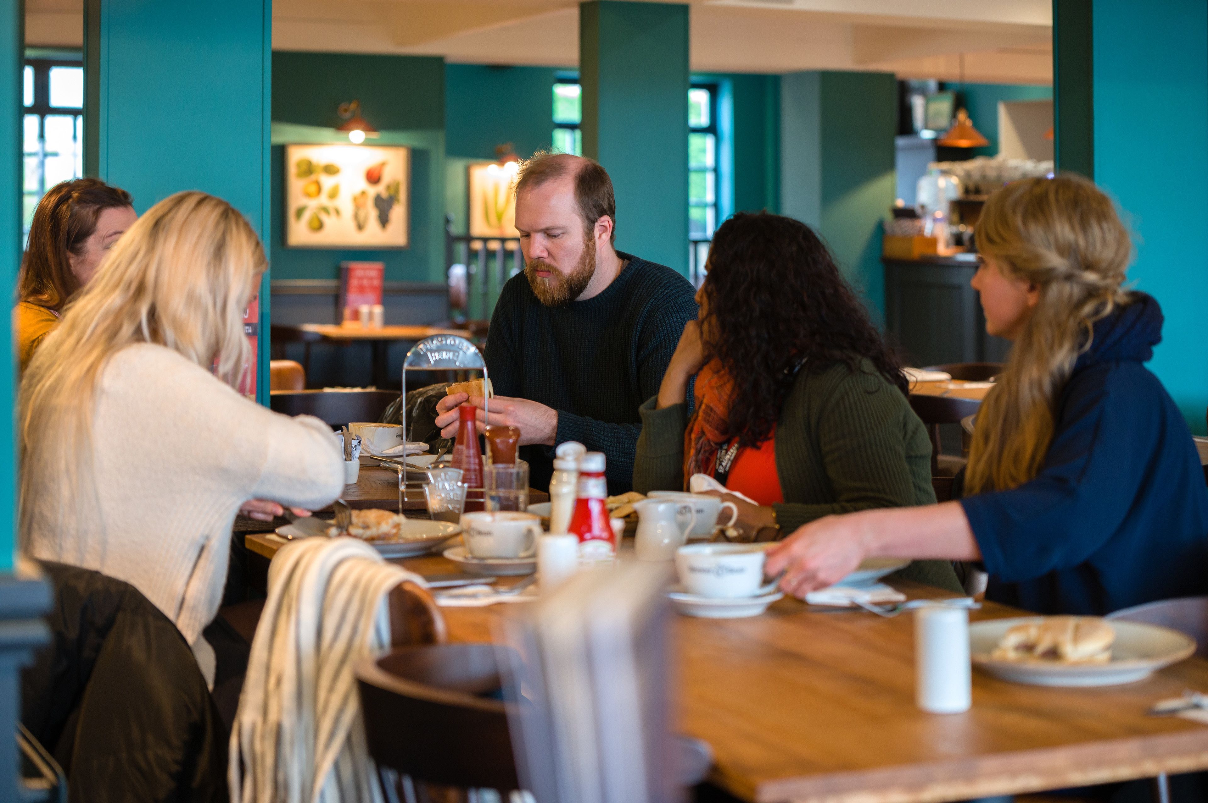 Eine Gruppe von Personen speist in einem Restaurant mit Holztisch, Stühlen und verschiedenen Speisen und Getränken.