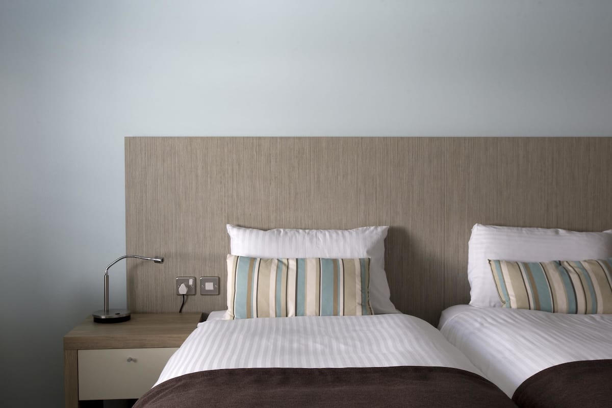 A hotel room with a wooden headboard, two beds with striped bedding, a bedside table with a lamp, and a drawer.