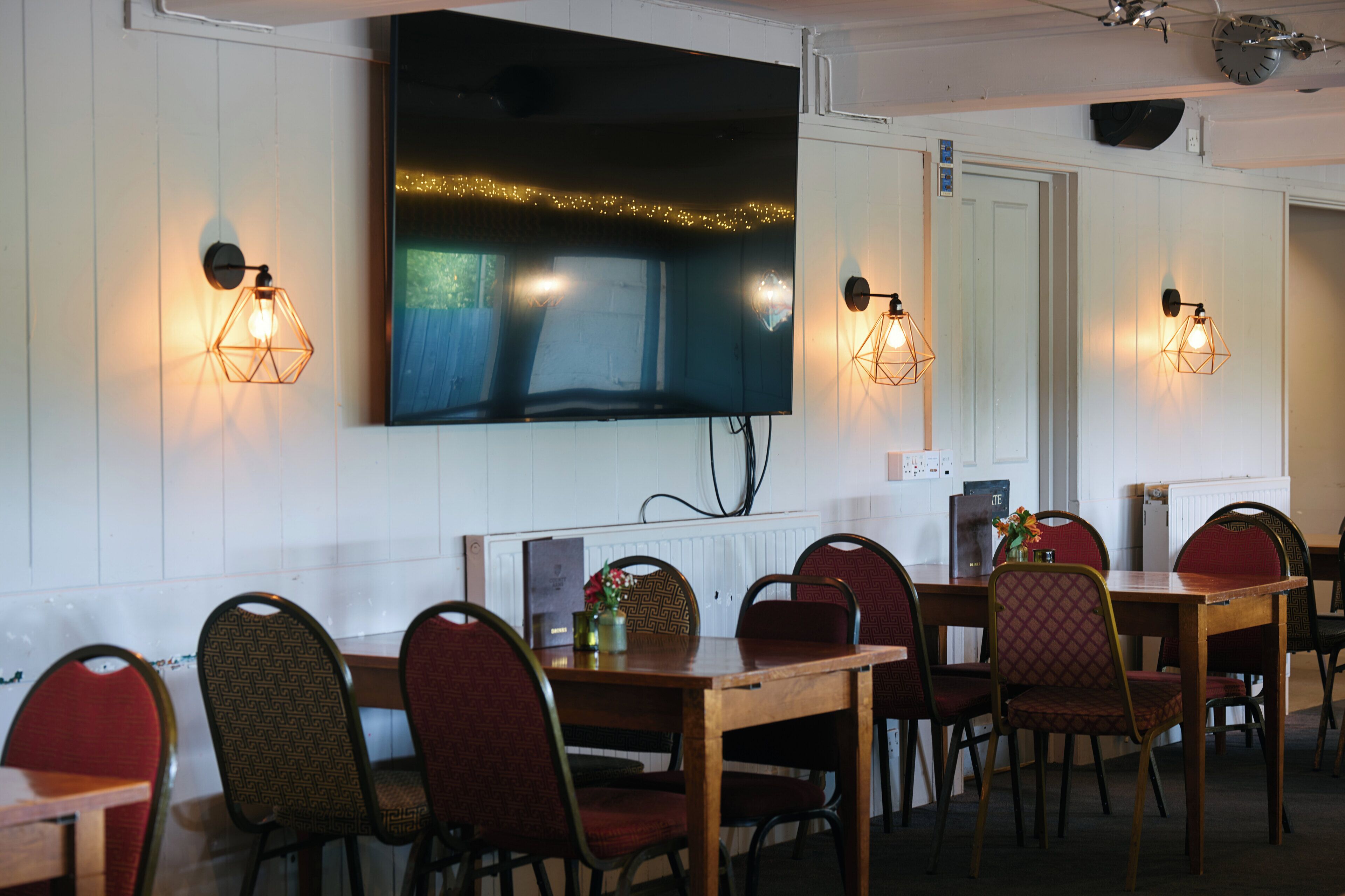 A dining area with tables and chairs, a flat-screen TV mounted on the wall, and wall-mounted lighting fixtures.