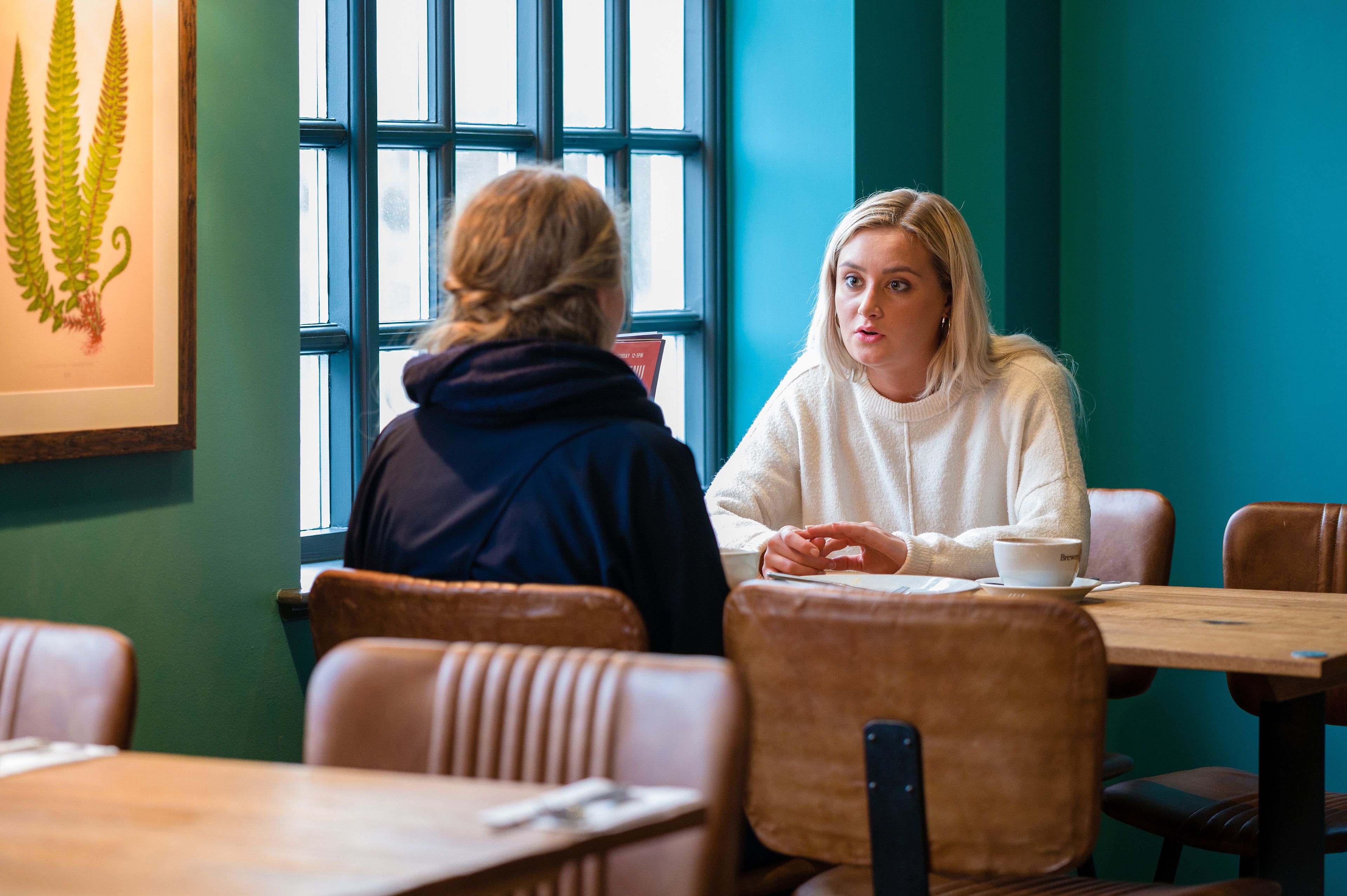 Dos personas sentadas en una mesa de madera en un café con paredes azul verdosas y una ventana con persianas.