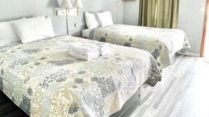 A hotel room with two beds, a hanging coat rack, a bedside table, a lamp, and a window with curtains. - Seacoast Motel (Wells)