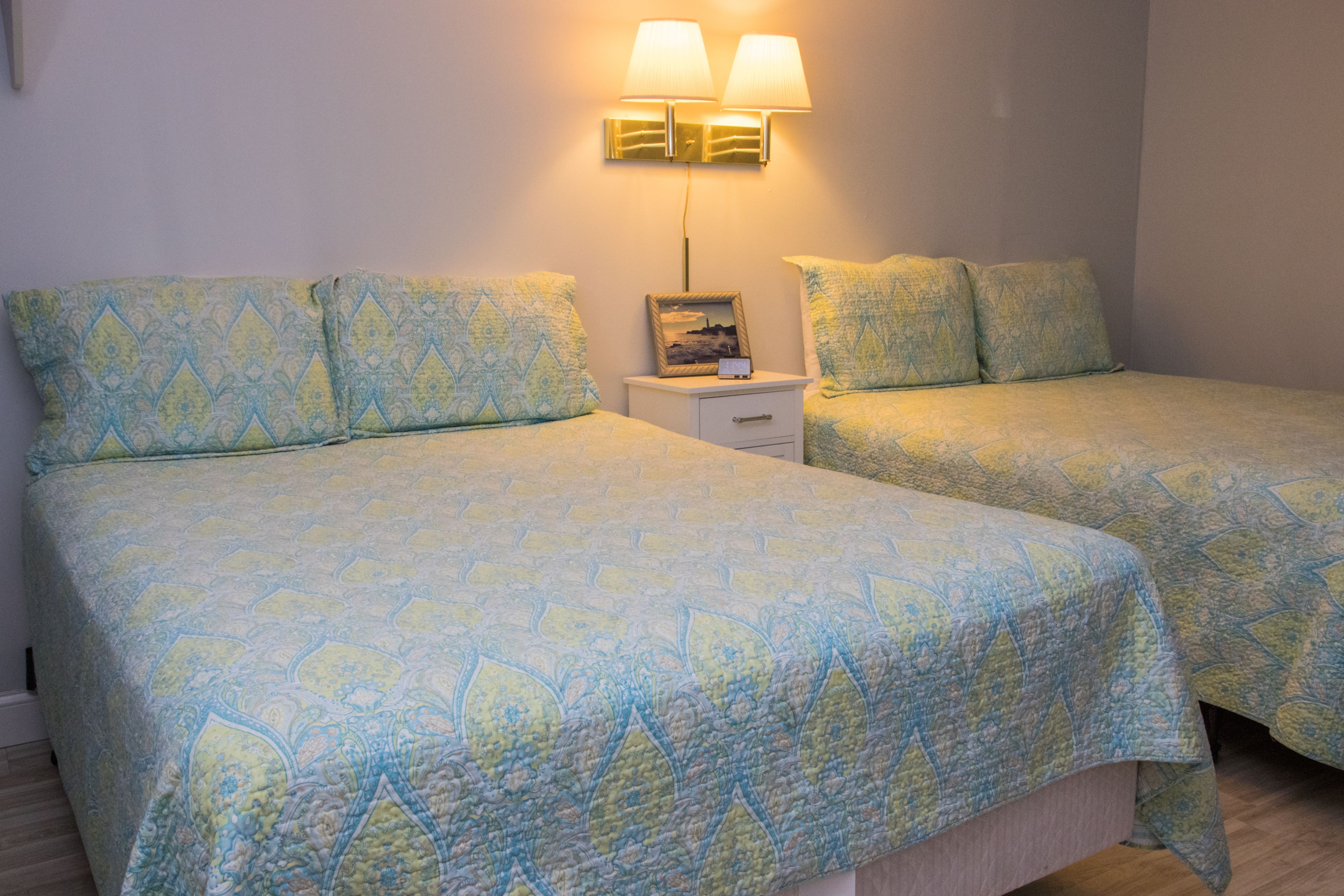 A room with two beds, each with patterned bedding, a bedside table with a lamp, and a framed picture on the wall.