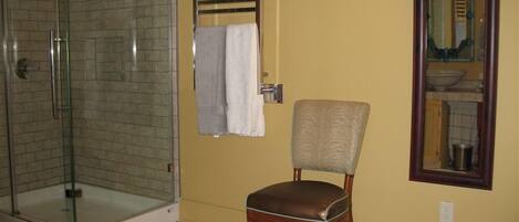 Une salle de bain avec une douche à parois de verre, une chaise en bois, un porte-serviettes et un miroir doté d’un cadre décoratif.