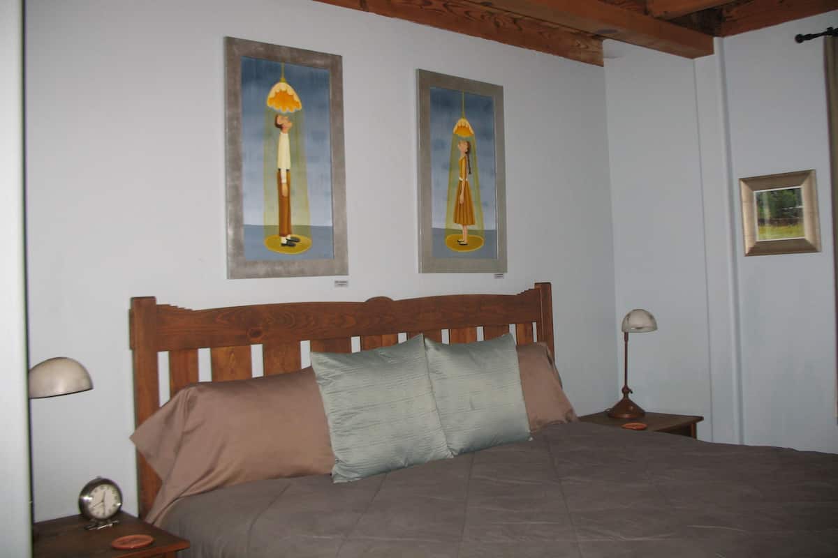 Un dormitorio con cabecera de madera, dos lámparas de noche, una cama con manta y almohadas marrones, y dos cuadros enmarcados en la pared.
