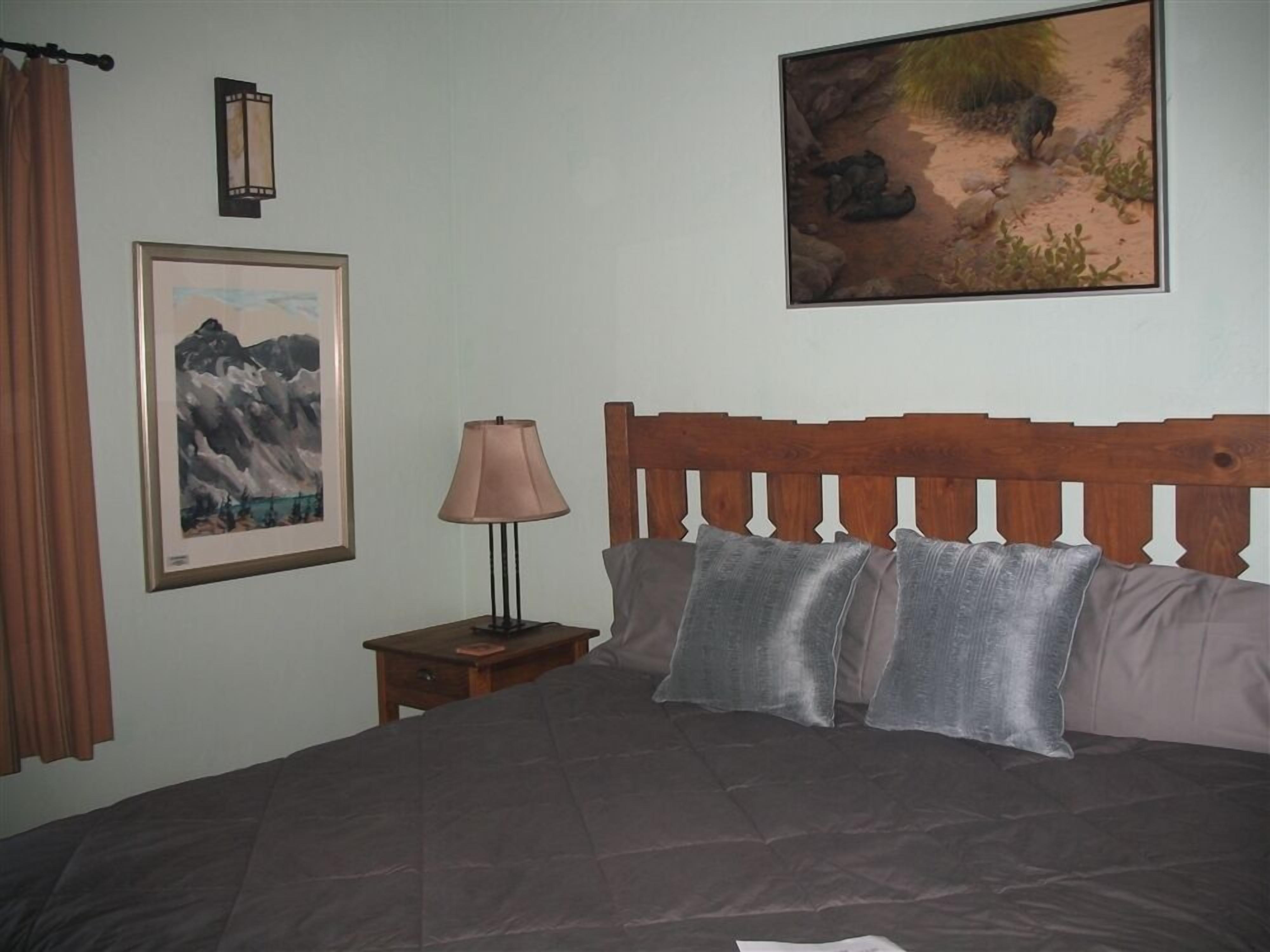 A bedroom with a wooden headboard, a bed with grey bedding, a nightstand with a lamp, and two framed pictures on the wall.