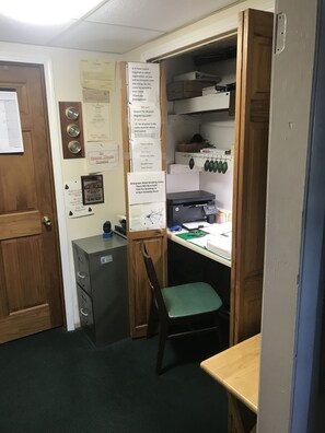 Een kleine kantoorruimte met een archiefkast, een bureau met een computer en een stoel. Op de muur hangen diverse berichten en borden, waaronder een bord dat persoonlijke cheques niet accepteert en een melding over het rookbeleid.