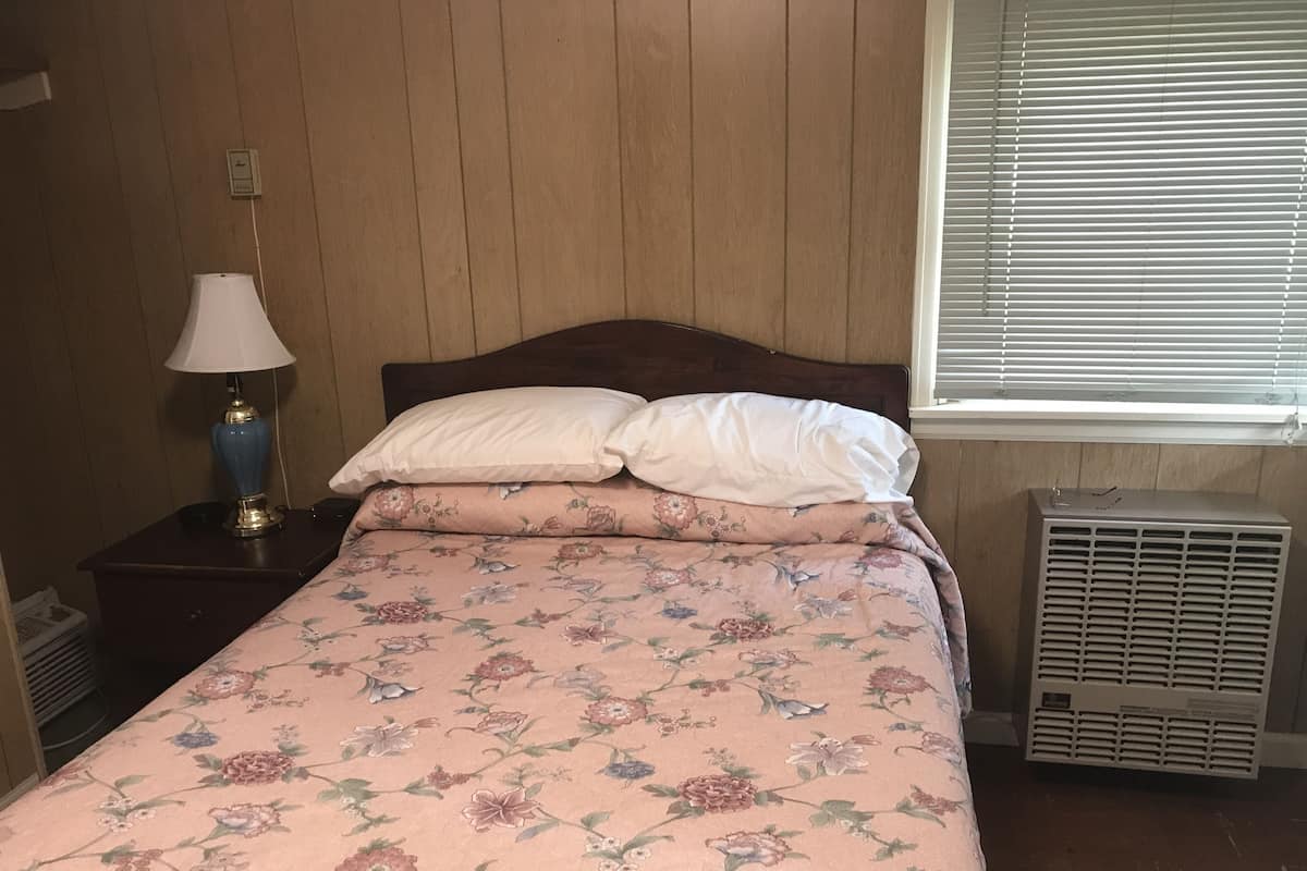 A bedroom with a wooden headboard, a floral bedspread, a bedside lamp, and a window with blinds.