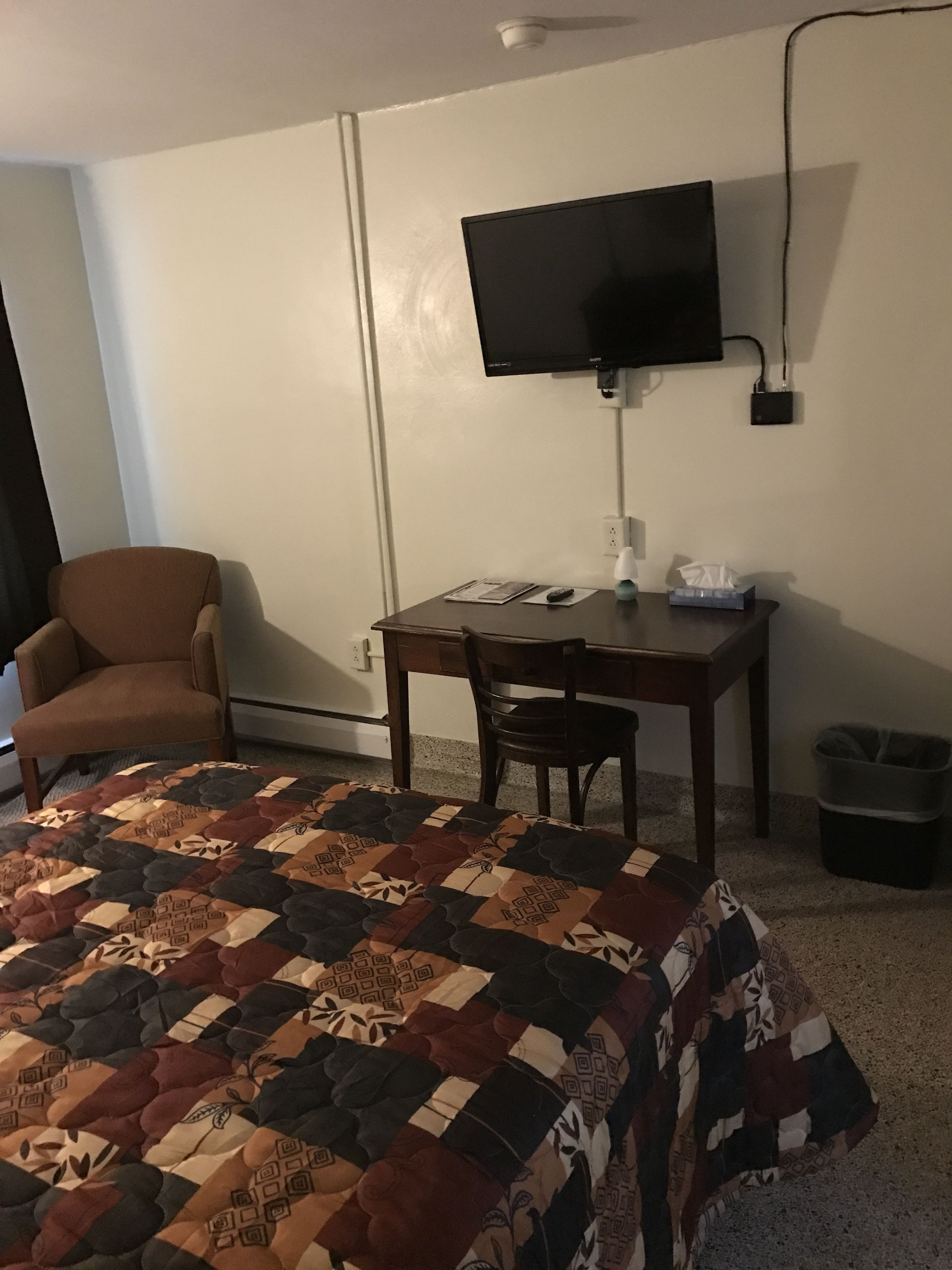 Una habitació d'hotel amb un llit, un escriptori, una cadira, un televisor fixat a la paret i un safata de recollida.