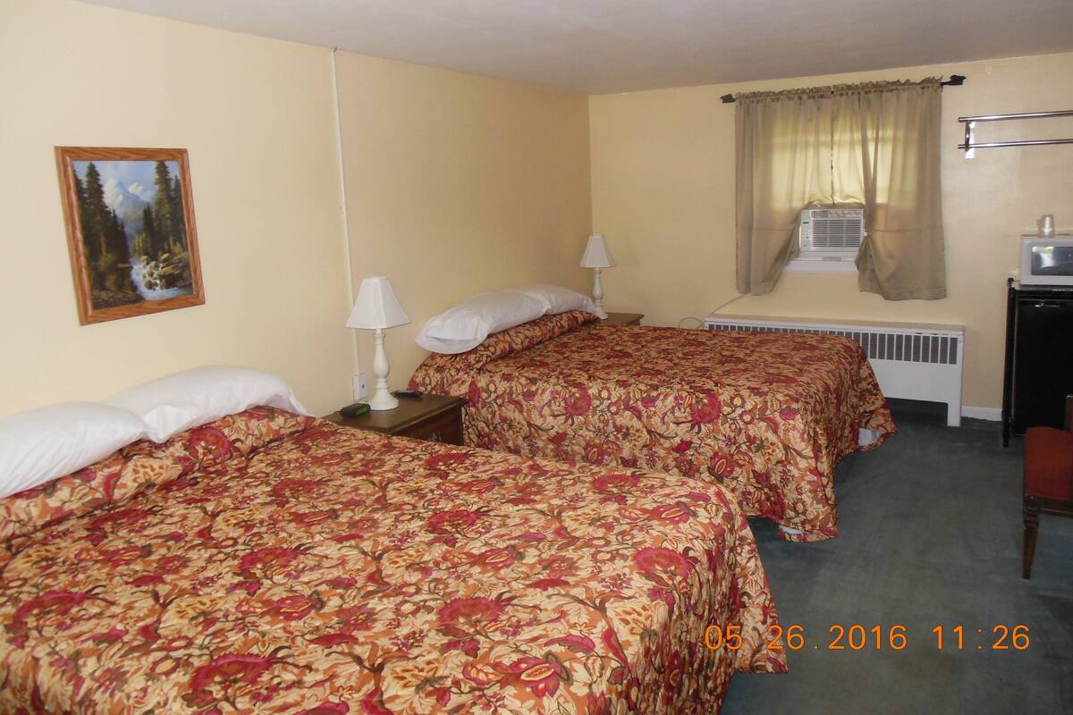 A hotel room with two beds, a painting, a window with curtains, an air conditioning unit, and a desk with a television.