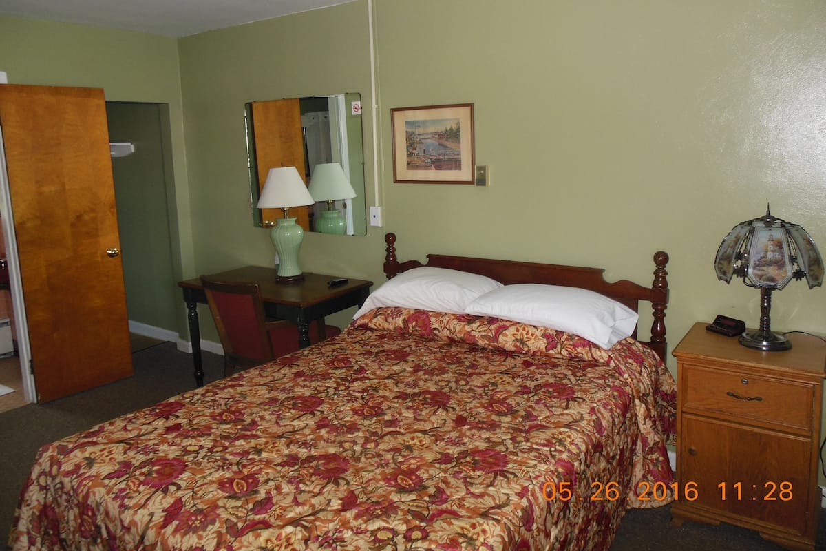 A hotel room with a bed, a desk, a chair, a nightstand, a lamp, a mirror, and a painting on the wall.