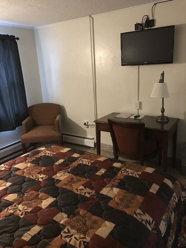 Una habitación de hotel con cama, escritorio, silla, televisor montado en la pared y ventana con cortinas.