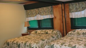 A bedroom with two beds, wooden headboard panels, green window blinds, and a patterned curtain. - The Evergreen Motel (Oswego)