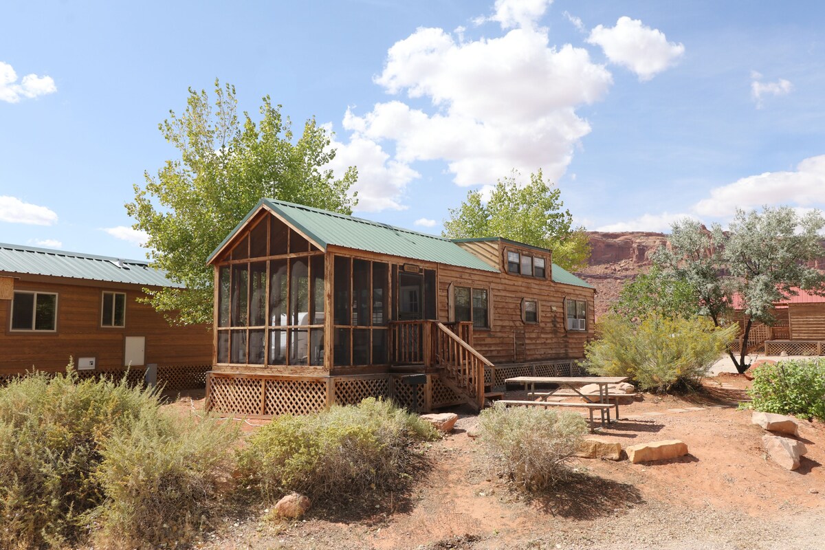A rustic cabin with a wooden exterior, a porch, and a picnic table in a desert setting.