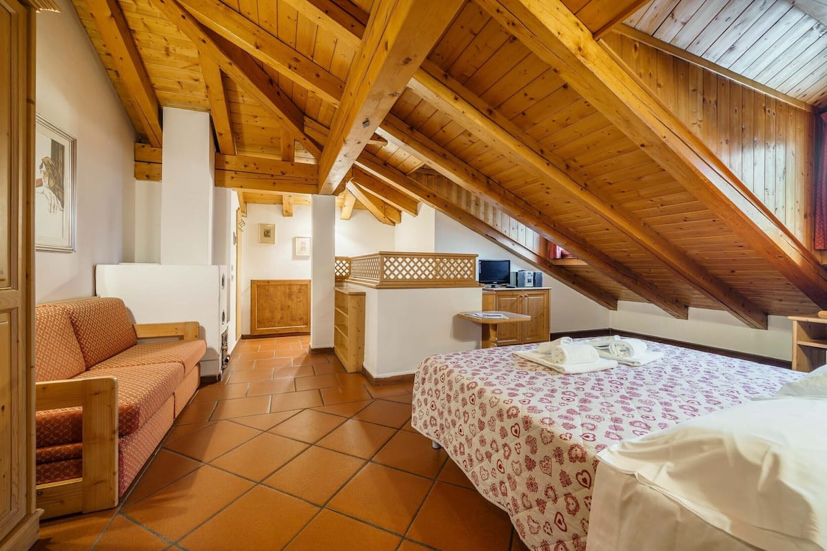 Een gezellig slaapkamer op zolder met een houten plafond, een bed met een bedsprei met dessin, een sofa en een klein bureau.