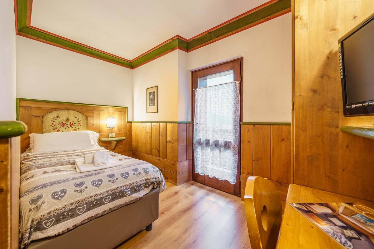 Hotelkamer met een houten bed, een tafeltje, een stoel, een televisie en een raam met gordijn.