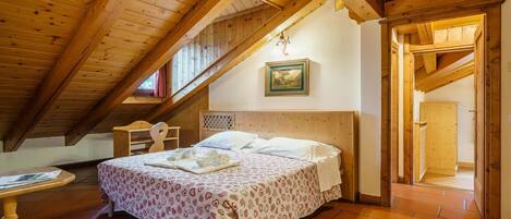 A cozy attic bedroom with a wooden ceiling, a bed with patterned bedding, a small table, and a framed picture on the wall.