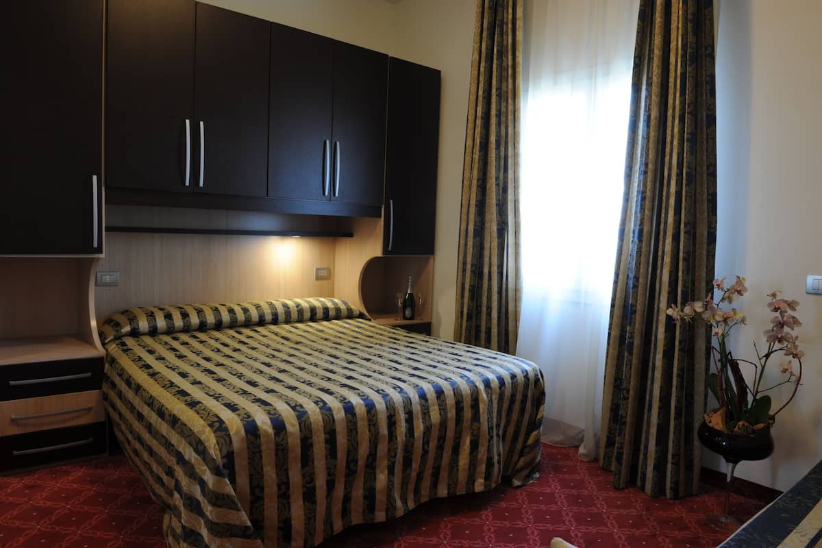 A hotel room with a bed, a nightstand, a dark wooden wardrobe, a window with curtains, and a potted orchid plant.