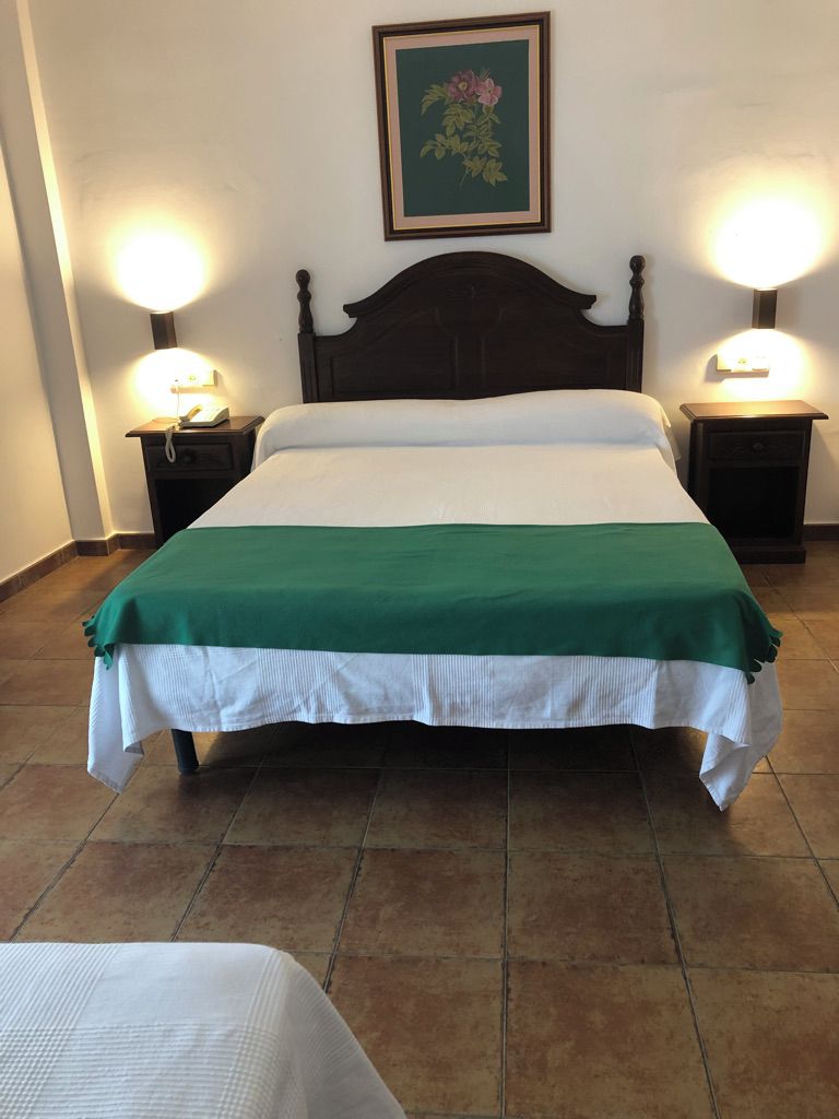 Una cama con una colcha verde y blanca, flanqueada por dos mesitas de noche con lámparas, en una habitación con suelo de baldosas.